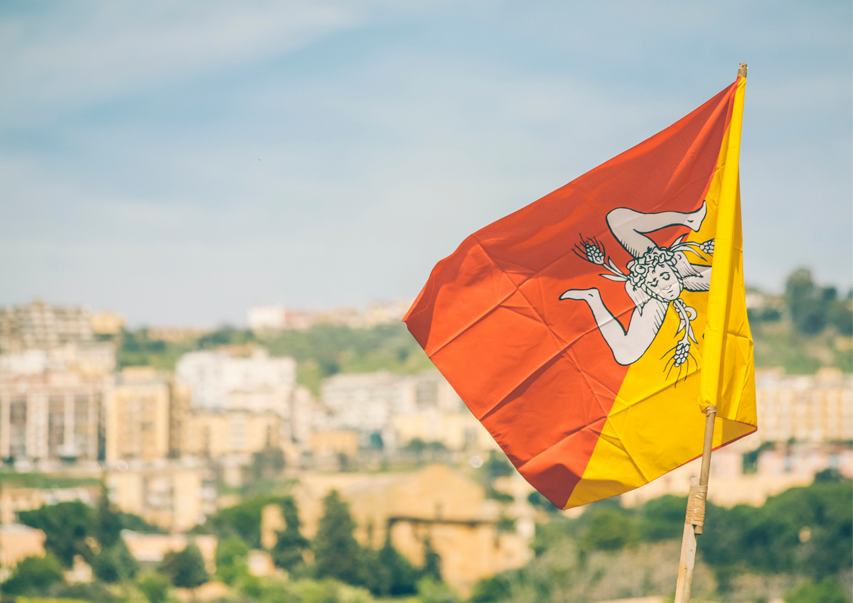 Bandiera della Sicilia con Trinacria al vento sopra un paesaggio urbano, perfetta per contenuti di marketing regionale e identità visiva isolana.